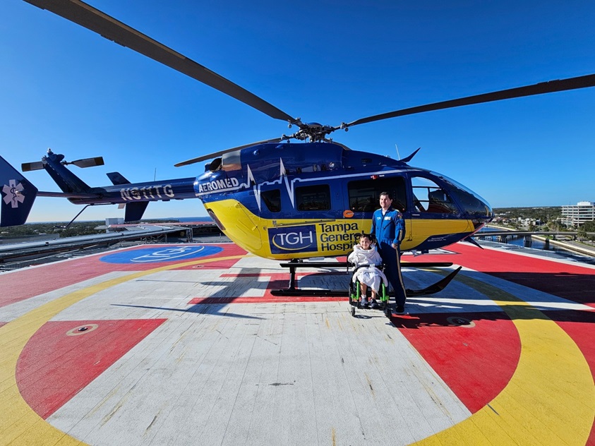 tgh pediatric patient in wheelchair and aeromed team member pose in front of tgh helicopter on the tgh helipad